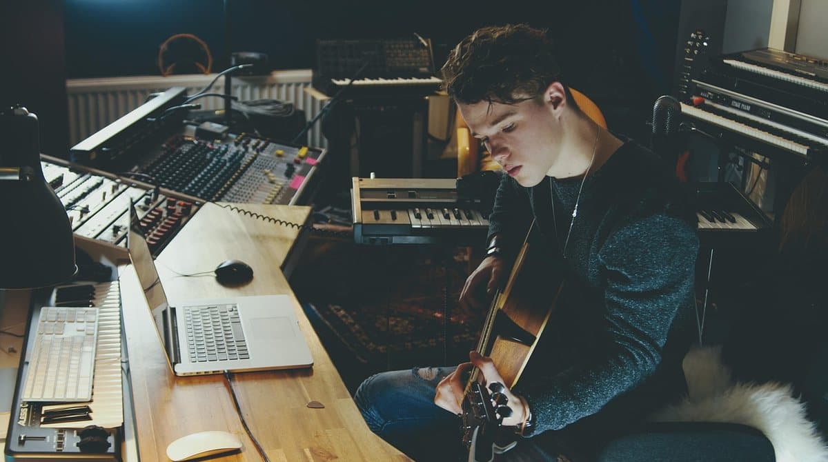 An independent musician playing guitar at a home studio desk with a laptop, mixing console, and keyboards